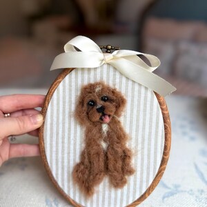 May include: An oval wooden embroidery hoop containing a needle-felted brown dog with a white chest patch and open mouth. The dog is mounted on beige and white striped fabric, and a cream-colored satin ribbon is tied at the top of the hoop.
