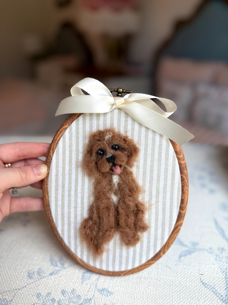 May include: An oval wooden embroidery hoop containing a needle-felted brown dog with a white chest patch and open mouth. The dog is mounted on beige and white striped fabric, and a cream-colored satin ribbon is tied at the top of the hoop.