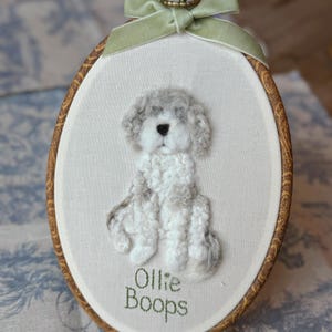 May include: An oval embroidery hoop containing a needle-felted white and gray dog on off-white fabric. The dog has a black nose and curly texture. Below the dog, the text "Ollie Boops" is stitched in green thread. A light green velvet ribbon is tied at the top.