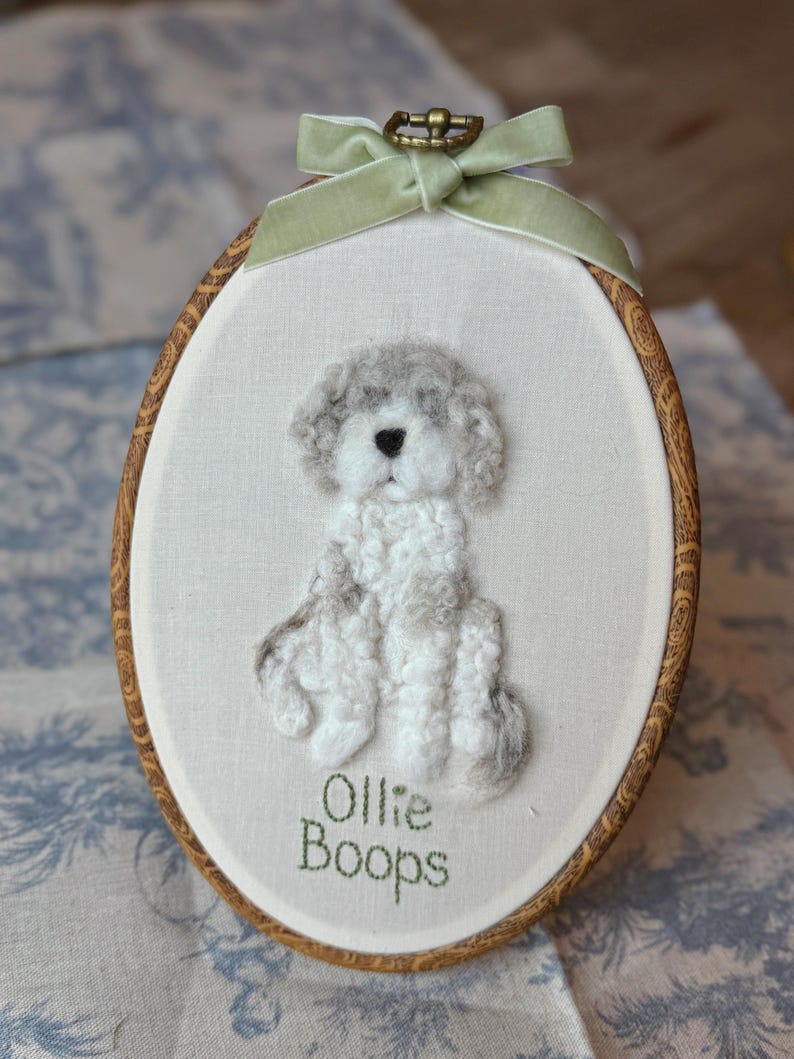 May include: An oval embroidery hoop containing a needle-felted white and gray dog on off-white fabric. The dog has a black nose and curly texture. Below the dog, the text "Ollie Boops" is stitched in green thread. A light green velvet ribbon is tied at the top.