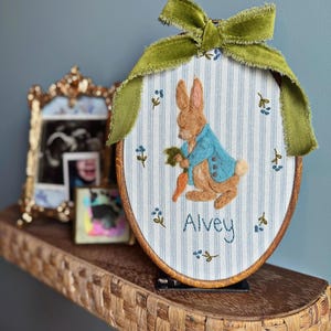 Puede incluir: Una obra de arte bordada de forma ovalada que representa un conejo con una chaqueta azul sosteniendo una zanahoria, con el nombre "Alvey" debajo. La obra está enmarcada en madera y adornada con un lazo de terciopelo verde. El fondo tiene rayas azules y blancas.