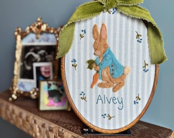 May include: An oval embroidery hoop featuring a brown rabbit wearing a blue jacket and holding a carrot, stitched onto white fabric with blue vertical stripes. The name Alvey is embroidered below the rabbit, accented by small blue flowers and a green velvet ribbon.
