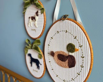 May include: Three oval embroidery hoops featuring needle-felted animal designs on white striped fabric. The largest hoop displays a mallard duck, while the two smaller hoops feature a brown and white dog and a black dog, each topped with a green velvet bow.