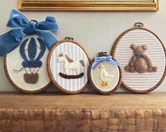 May include: Four circular wooden embroidery hoops featuring needle-felted designs: a blue and white hot air balloon with a velvet bow, a white rocking horse, a small white duck with a blue velvet bow, and a brown teddy bear with a blue bow.