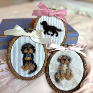 May include: Three oval wooden embroidery hoops containing needle-felted dog portraits on fabric. One hoop features a black and tan dog with a blue bow, one a black dog, and one a grey and tan dog with a pink bow, each topped with a satin ribbon.
