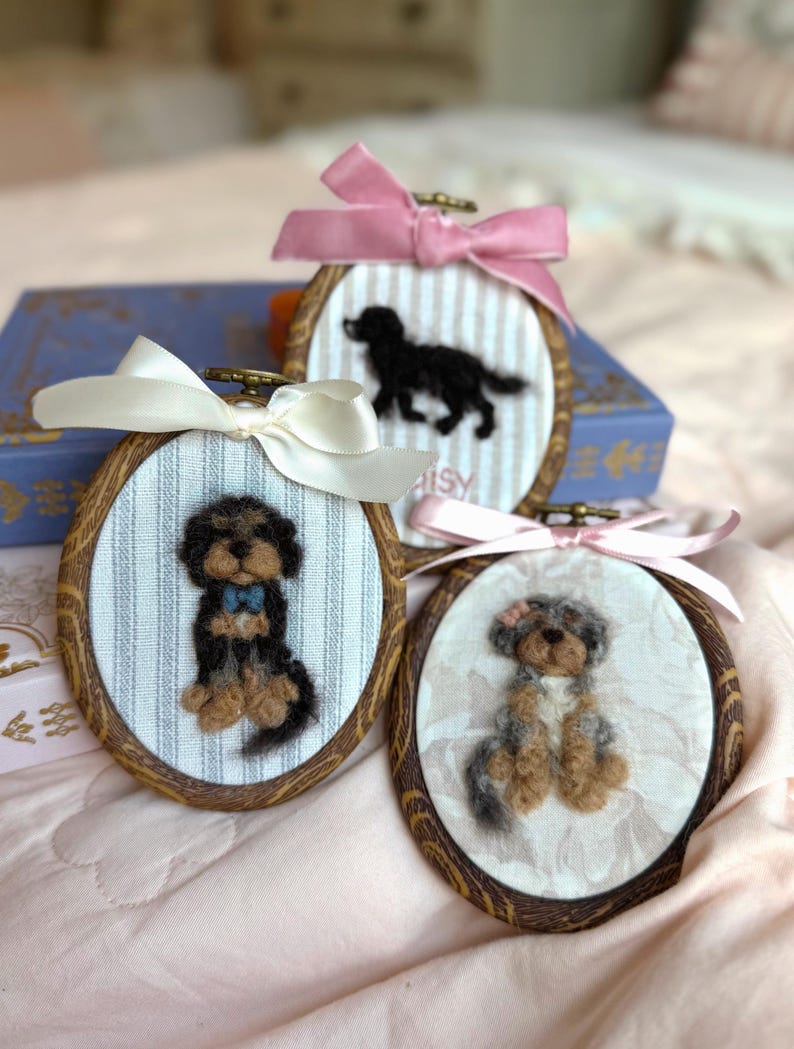 May include: Three oval wooden embroidery hoops containing needle-felted dog portraits on fabric. One hoop features a black and tan dog with a blue bow, one a black dog, and one a grey and tan dog with a pink bow, each topped with a satin ribbon.