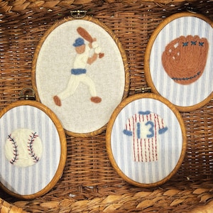 May include: Four baseball-themed embroidery hoops in a woven basket. The hoops feature a baseball, a glove, a jersey with the number 13, and a baseball player. The backgrounds are either striped or solid.
