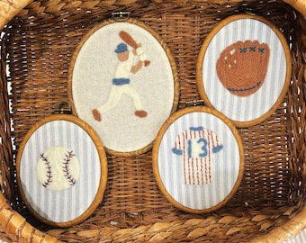 May include: Four baseball-themed embroidery hoops in a woven basket. The hoops feature a baseball, a glove, a jersey with the number 13, and a baseball player. The backgrounds are either striped or solid.