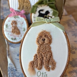 May include: An oval wooden embroidery hoop containing a needle-felted brown curly-haired dog portrait on white fabric. The name Otis is embroidered in green thread below the dog. A light green velvet ribbon is tied at the top of the hoop.