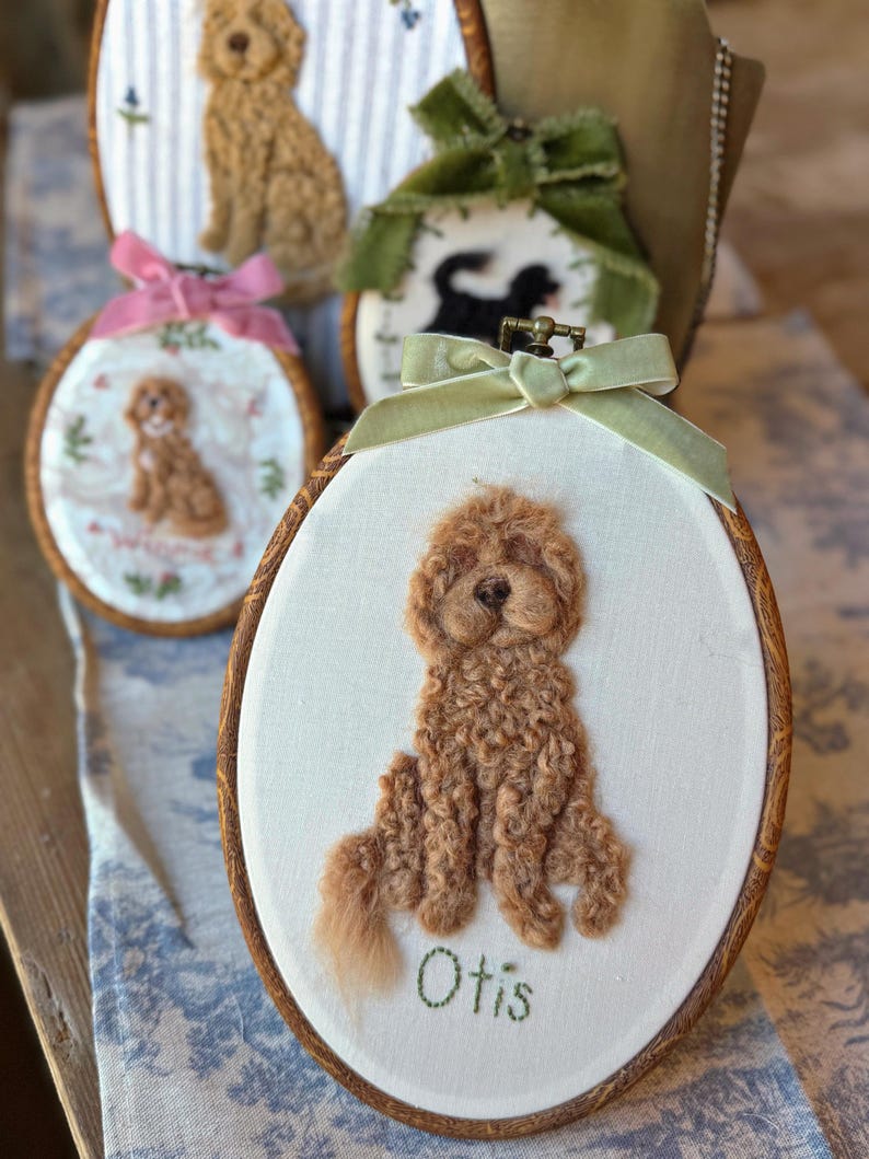 May include: An oval wooden embroidery hoop containing a needle-felted brown curly-haired dog portrait on white fabric. The name Otis is embroidered in green thread below the dog. A light green velvet ribbon is tied at the top of the hoop.