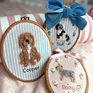 May include: Three oval wooden embroidery hoops featuring needle-felted dog portraits. One hoop shows a brown dog named Cooper on striped fabric. Another features a black and white dog named Blue with a blue velvet bow. A third shows a small dog named Daisy surrounded by embroidered daisies.