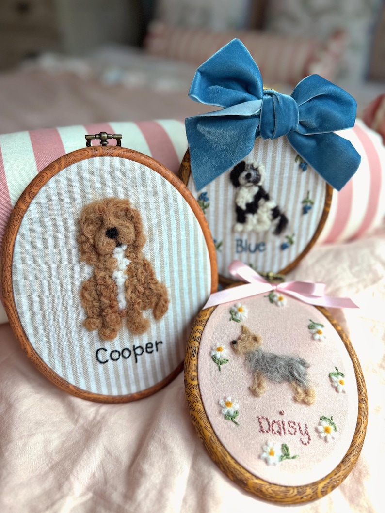 May include: Three oval wooden embroidery hoops featuring needle-felted dog portraits. One hoop shows a brown dog named Cooper on striped fabric. Another features a black and white dog named Blue with a blue velvet bow. A third shows a small dog named Daisy surrounded by embroidered daisies.