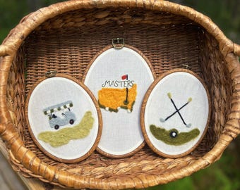 May include: Three oval embroidery hoops featuring golf-themed designs on white fabric. One hoop shows a light blue golf cart on a green patch, another displays a yellow map of the United States with the text MASTERS and a red flag, and the third shows crossed golf clubs over a green patch.