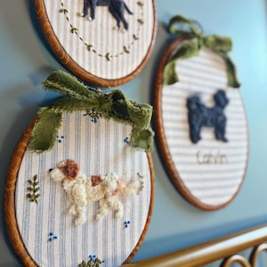 May include: Three embroidered dog portraits in wooden hoops. Each features a different dog breed, with details like fur texture and color variations. The backgrounds are striped fabric, and the hoops are adorned with green ribbon.