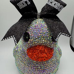 May include: A cheerleading bow with a sparkling black glitter finish and the words "HIT ZERO" in white. The bow is adorned with iridescent rhinestones and a large, bright orange nose.