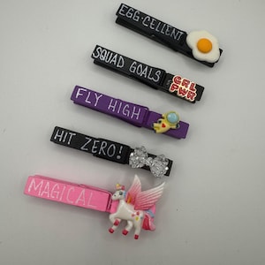Può includere: Una collezione di mollette decorative con vari disegni e testo. Le mollette sono nere, viola e rosa, con frasi come "EGG-CELLENT" e "MAGICAL". Ogni molletta presenta un fascino unico, tra cui un uovo fritto, un unicorno e un fiocco.