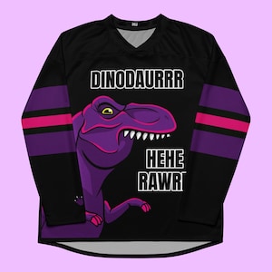 May include: A black long-sleeve jersey featuring a purple dinosaur graphic with the text DINODAURRR HEHE RAWR. The sleeves have horizontal purple and pink stripes. The shirt has a V-neck collar and a black base color with a graphic print on the front.