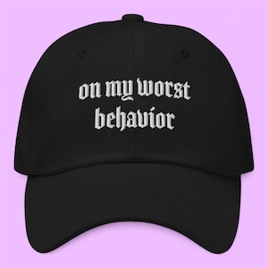 May include: A black baseball cap featuring the text "on my worst behavior" embroidered in white thread across the front. The cap has a curved brim, a structured crown with a button on top, and eyelets for ventilation, creating a simple design.
