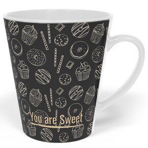 May include: White ceramic latte mug featuring a black background adorned with a cream-coloured pattern of donuts, cupcakes, and cookies. The words "You are Sweet" are printed on the front in gold.