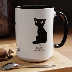 May include: White ceramic mug with a black interior and handle. The mug features a black cartoon cat with an angry expression and the text "Cat. Coffee. Boundaries.". Paw prints are also visible on the mug.