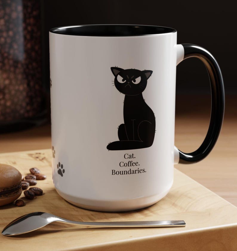 May include: White ceramic mug with a black interior and handle. The mug features a black cartoon cat with an angry expression and the text "Cat. Coffee. Boundaries.". Paw prints are also visible on the mug.