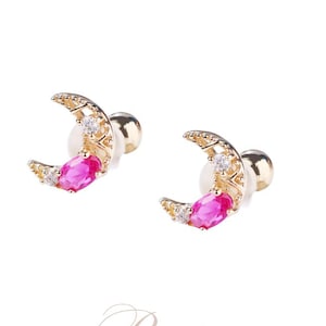 May include: Gold-toned crescent moon earrings with a vibrant pink gemstone center and small white accent stones. The earrings have a post-back closure. The image also includes the word "Rivita" in cursive script.