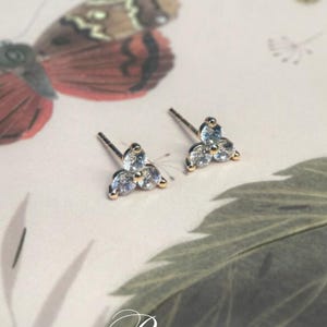 May include: A pair of stud earrings with a three-stone design. Each earring has three clear, round-cut stones set in gold-tone metal. The earrings are displayed on a patterned surface with a butterfly and leaf.