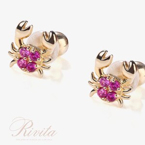 May include: A pair of gold-colored crab-shaped earrings. Each crab features a cluster of pink gemstones at the center and has detailed claws. The earrings are set against a light background.