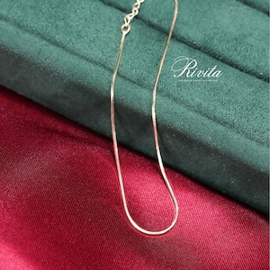 May include: A delicate silver chain necklace with a snake chain design. The necklace is displayed on a red satin surface and a green velvet surface. The word "Rivita" is written in cursive on the green surface.