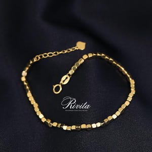 May include: A gold bracelet with square beads and a delicate chain. The bracelet has a heart-shaped charm and a clasp closure. The bracelet is displayed on a dark blue background. The word "Rivita" is written in cursive.