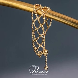 Może przedstawiać: Delikatny, złoty pierścionek z łańcuszkiem o unikalnym, siatkowym wzorze, zawieszony na złotym pręcie. Pierścionek posiada zwisający łańcuszek z kulistą koralikiem na końcu. Obraz zawiera tekst "Rivita" w eleganckiej czcionce.