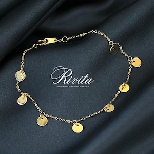 May include: A delicate gold anklet featuring small, round disc charms evenly spaced along a fine chain. The anklet is displayed on a dark, textured surface. The word "Rivita" is displayed in cursive font.