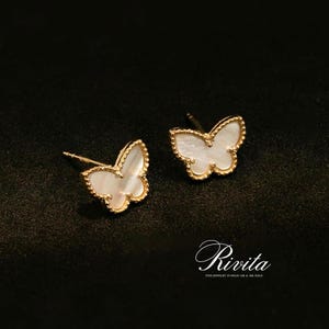 Op de afbeelding: Een paar gouden vlinder-oorstekers met een witte parelmoer inleg. De oorbellen hebben een gouden, kralenrand en zijn geplaatst tegen een donkere achtergrond. De merknaam "Rivita" wordt cursief weergegeven.