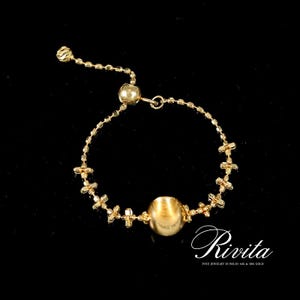 Pode incluir: Uma pulseira de ouro ajustável com uma grande conta central e delicados detalhes em forma de cruz. A pulseira apresenta uma pequena corrente e um fecho para ajustar o tamanho. A marca "Rivita" é exibida em escrita cursiva.