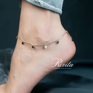 May include: A gold anklet with small, dangling charms. The anklet features a delicate chain with alternating black and gold circular charms. The word "Rivita" is displayed in cursive script. Fine jewelry in solid 14k & 18k gold.