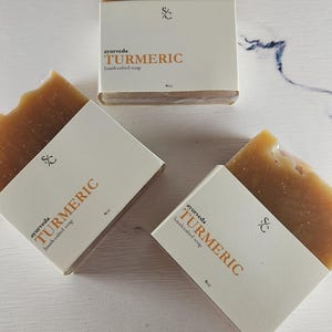 May include: Three bars of turmeric soap, each wrapped in white paper with the text "ayurveda TURMERIC handcrafted soap 4oz".