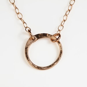 May include: A copper-colored necklace featuring a hammered, open circle pendant. The pendant is connected to a delicate chain with oval links. The necklace is displayed against a white background, highlighting the warm tones of the metal.