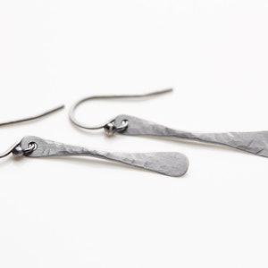 May include: A pair of silver dangle earrings with a hammered texture. The earrings are long and thin, with a slightly curved shape.