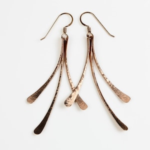 May include: A pair of copper-colored dangle earrings. Each earring features a long, hammered metal design with a curved hook for the ear. The earrings have a simple, elegant design, with a textured finish. The earrings are displayed on a white background.