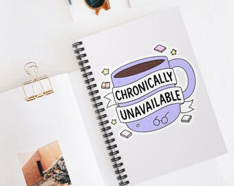 May include: A sticker featuring a light purple coffee mug with the words "Chronically Unavailable" on a banner. The mug is filled with brown liquid and surrounded by small stars, books, and glasses. The sticker is on a white spiral notebook.