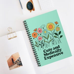 May include: A spiral-bound notebook with a light teal cover featuring a floral design in orange, pink, yellow, and white. The text "Cute and Medically Expensive" is printed in black. The notebook is open on a white surface.