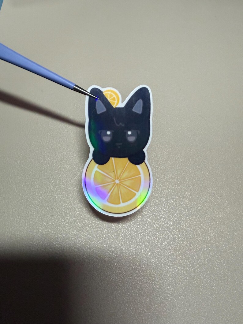 Op de afbeelding: Een sticker met een zwarte kat met een citroen schijf ontwerp. De kat kijkt over een gele en oranje citroen schijf met een holografische rand. Een kleine citroen schijf zit op het hoofd van de kat.