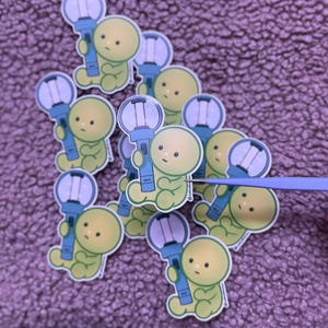 May include: A collection of stickers featuring a cartoon character holding a blue and white light stick. The character is yellow-green with a simple design. The stickers are on a purple textured surface.