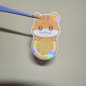 Op de afbeelding: Een sticker met een cartoon hamster met oranje vacht, die een citroen schijf vasthoudt. De sticker heeft een holografisch effect en wordt vastgehouden door een pincet. De hamster heeft een citroen schijf op zijn hoofd.