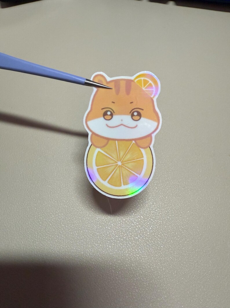 Op de afbeelding: Een sticker met een cartoon hamster met oranje vacht, die een citroen schijf vasthoudt. De sticker heeft een holografisch effect en wordt vastgehouden door een pincet. De hamster heeft een citroen schijf op zijn hoofd.