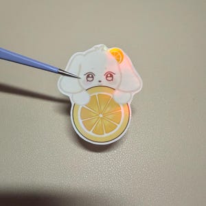 Op de afbeelding: Een witte sticker in de vorm van een konijn met een citroen. Het konijn heeft grote ogen en houdt de citroen vast. De sticker zit op een lichtgekleurd oppervlak. Een gereedschap wijst naar de sticker.