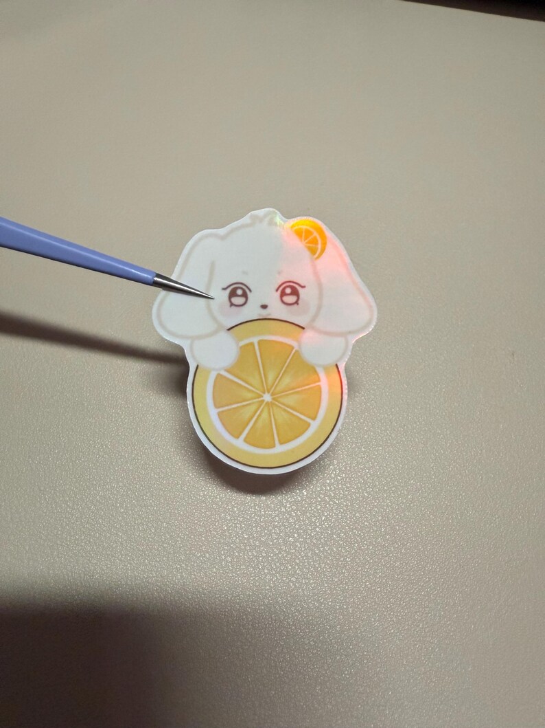 Op de afbeelding: Een witte sticker in de vorm van een konijn met een citroen. Het konijn heeft grote ogen en houdt de citroen vast. De sticker zit op een lichtgekleurd oppervlak. Een gereedschap wijst naar de sticker.