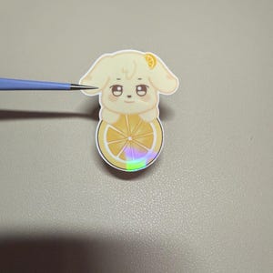 Op de afbeelding: Een gestanste sticker met een cartoonhond met een citroenschijfje als lichaam. De hond is lichtgeel met een klein citroenschijfje op zijn hoofd. Het citroenschijfje lichaam heeft een holografisch effect. De sticker ligt op een grijze ondergrond.