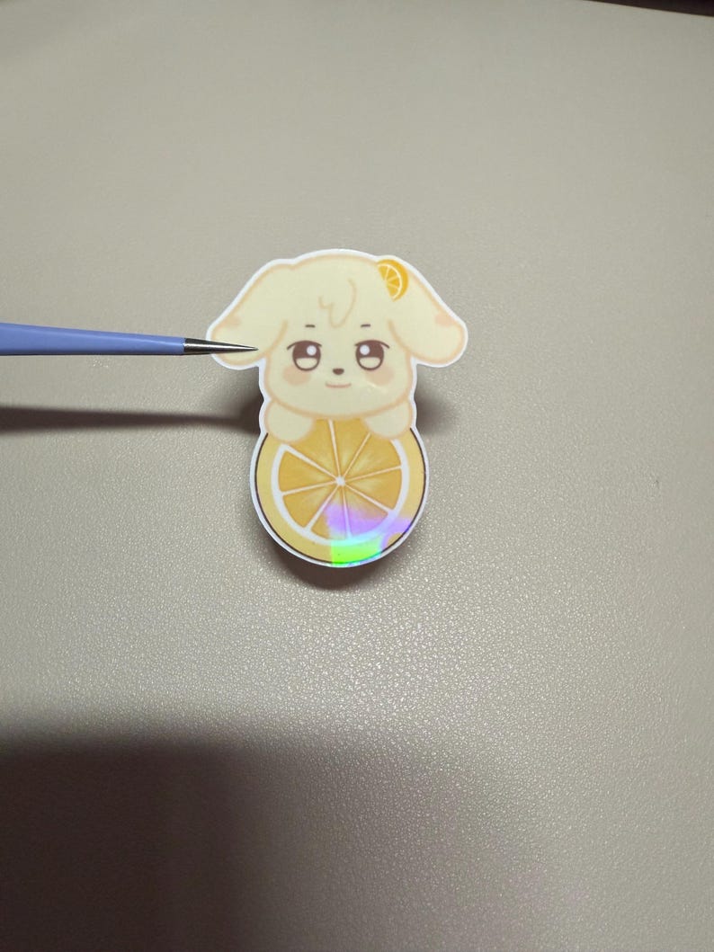 Op de afbeelding: Een gestanste sticker met een cartoonhond met een citroenschijfje als lichaam. De hond is lichtgeel met een klein citroenschijfje op zijn hoofd. Het citroenschijfje lichaam heeft een holografisch effect. De sticker ligt op een grijze ondergrond.