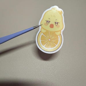 Op de afbeelding: Een gele sticker met een cartoon eend karakter met een sinaasappelschijf. De eend heeft een chagrijnige uitdrukking en houdt de sinaasappelschijf vast. De sticker wordt vastgehouden door een blauw en zilverkleurig gereedschap.